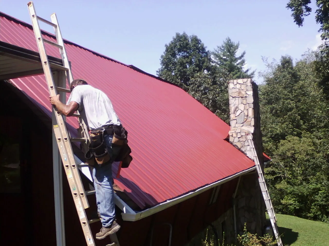 Roofer on a ladder beside a red metal roof and stone chimney — Soffit & Fascia Repair by Roofing Brothers in Point Marion